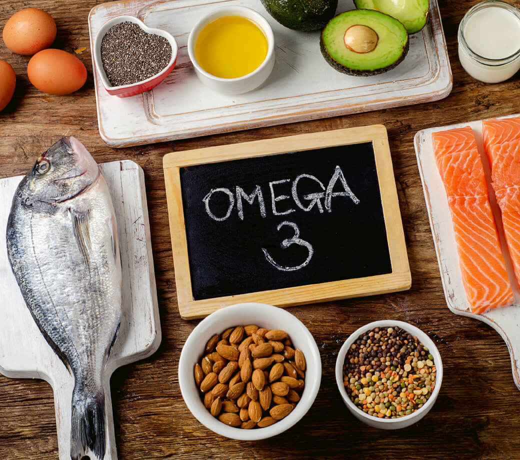 Natural Pharmaceuticals OmegaMarine™ Premium Omega-3 rasvhapete rikkad toidud nagu kala, lõhe, avokaado, mandlid, chia seemned ja munad puidust lauapinnal.